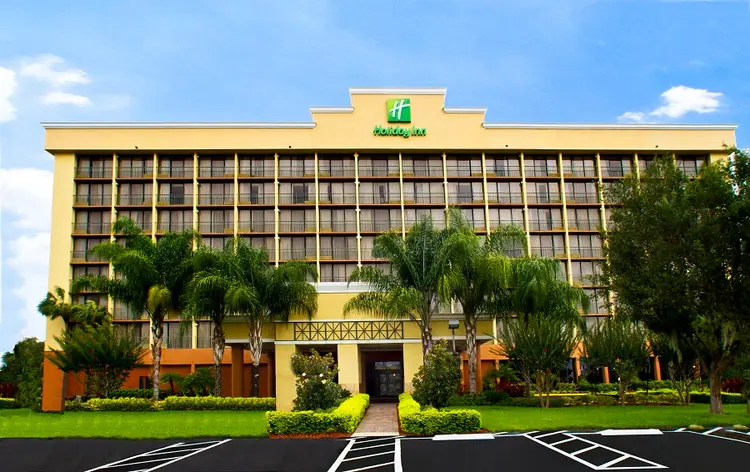 Exterior of the Holiday Inn with hotel signage and a manicured lawn with palm trees, hedges and a brick pathway