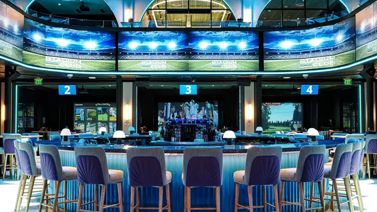 A bar with a round counter and padded stools surrounded by video screens
