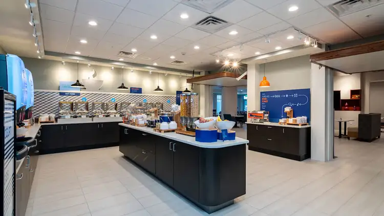 A large room with an island in the middle and counters filled with fruit, cereal and a row of hot dishes