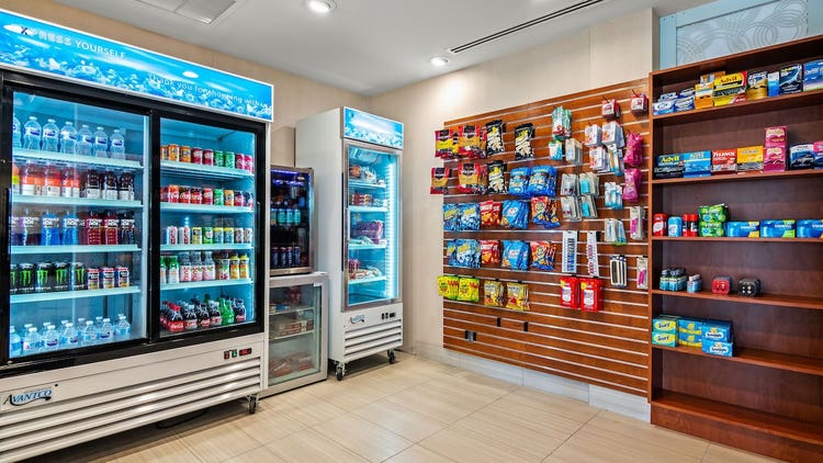 Three refrigerators with glass doors revealing waters, juices and soft drinks, hanging packages of candy and crackers, and shelves that hold pain relievers, fabric softener sheets and more