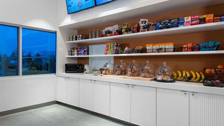 A room with a microwave, stocked with bagels, sweet rolls, muffins, bananas, oranges, protein bars, pain relievers and more