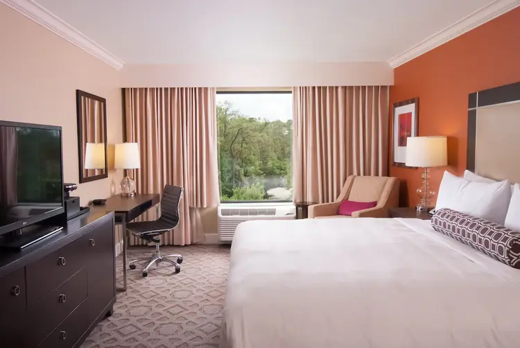 A guest room featuring a dresser, TV, desk, chair, lamp, mirror, large window and drapes, an upholstered chair, a side table with lamp and a bed