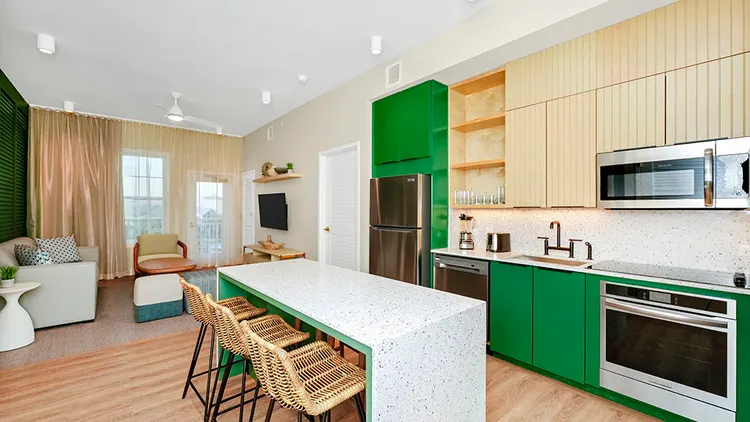 A hotel suite kitchen at Embassy Suites by Hilton Orlando Sunset Walk featuring a kitchen island, bar stools, an oven, a sink, a dishwashing machine, a refrigerator, a sofa, a coffee table and a TV