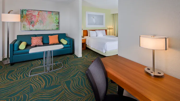 A sofa with 2 tray stands near a king bed with 2 night stands across from a desk with a chair