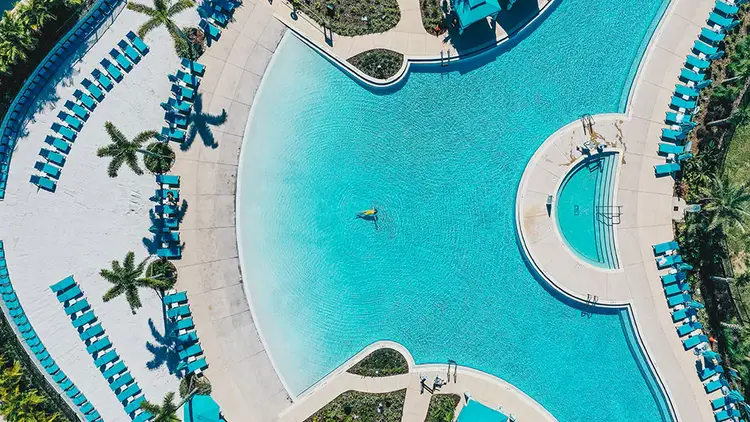 A woman floats in the middle of a large pool with nearby seating areas and palm trees