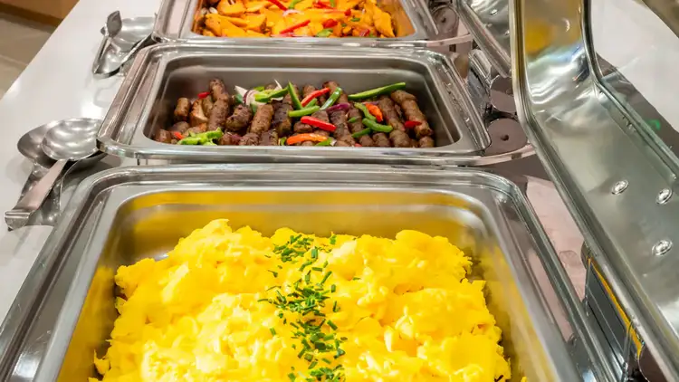 A breakfast buffet station with chafing dishes filled with eggs, sausage and roasted potatoes