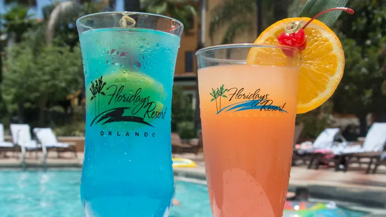 Two cocktails in glasses garnished with fruit on a table by a swimming pool