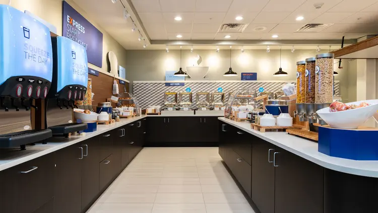 A large room with an island in the middle and counters filled with fruit juice dispensers, fruit, cereal and a row of hot dishes