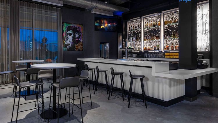 A modern bar with bar stools and tall tables and chairs, a painting of a man on the wall and cabinets holding liquor bottles